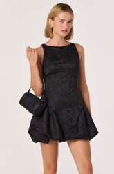 A woman is wearing a sleeveless, knee-length black dress with a subtle floral pattern. The dress has a fitted bodice and a voluminous, puffed skirt with a bubble hem. She accessorizes with bold, geometric gold earrings and a small black shoulder bag.